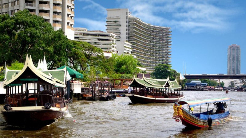 A turbulent river with Thai riverboats and hotel buildings on its banks