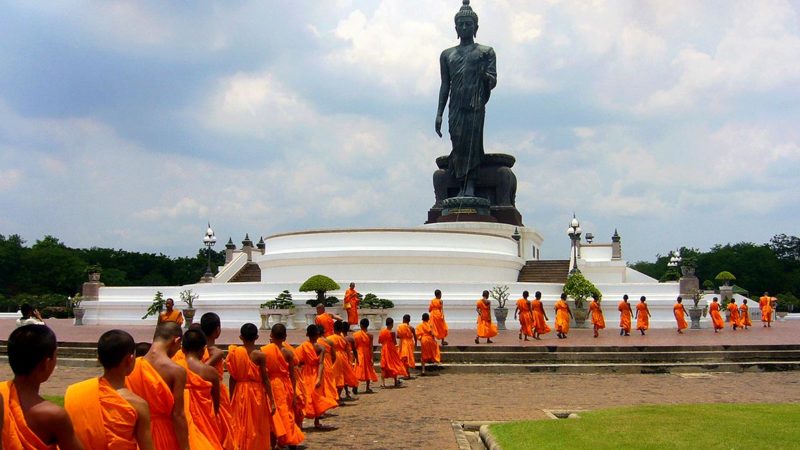 A large bronze statue of the walking buddha with a line of buddhist monks in robes walking towards it