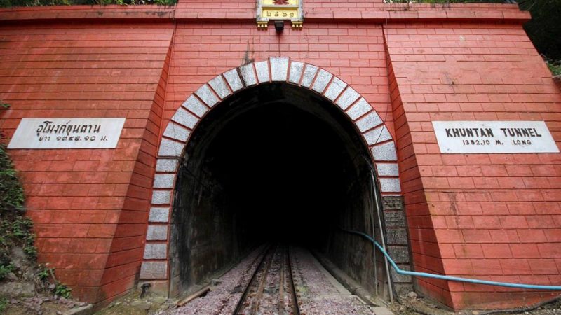 An arched entrance to a dark railway tunnel lined with red bricks and an ornate Thai sign